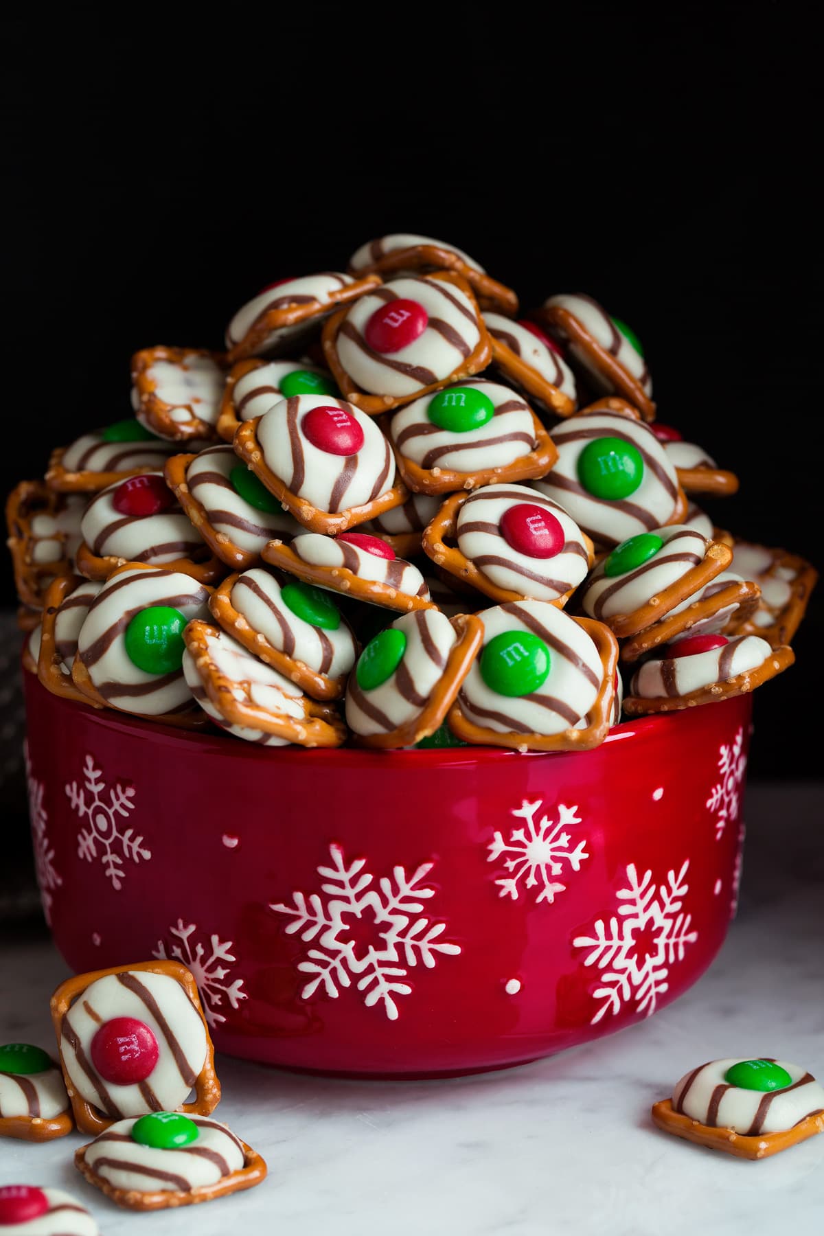 Pretzel M&M Hugs in a red snowflake bowl. The consist of square pretzel base, hug chocolate and red and green M&M's.