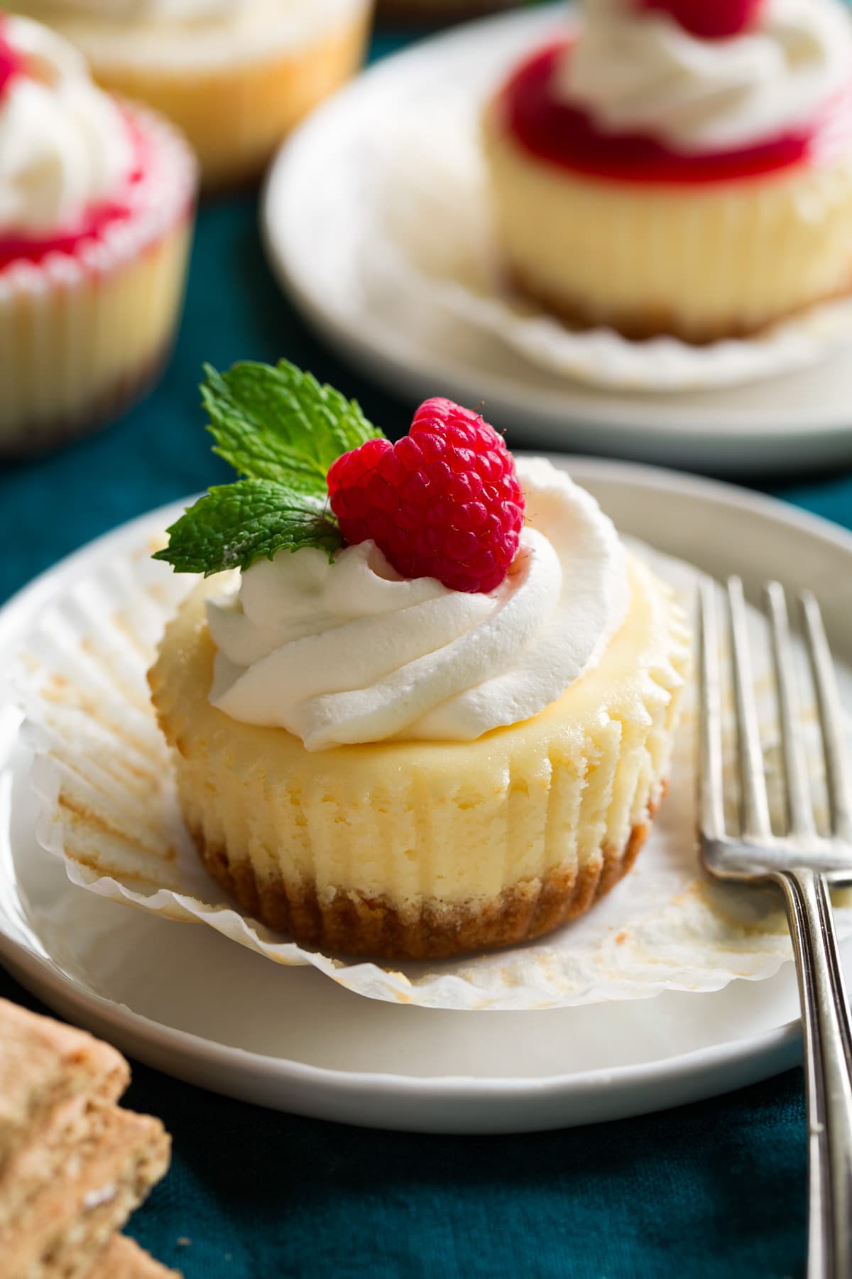 Mini cheesecake shown on a dessert plate unwrapped from cupcake liner. It is topped with whipped cream, fresh raspberry and mint.