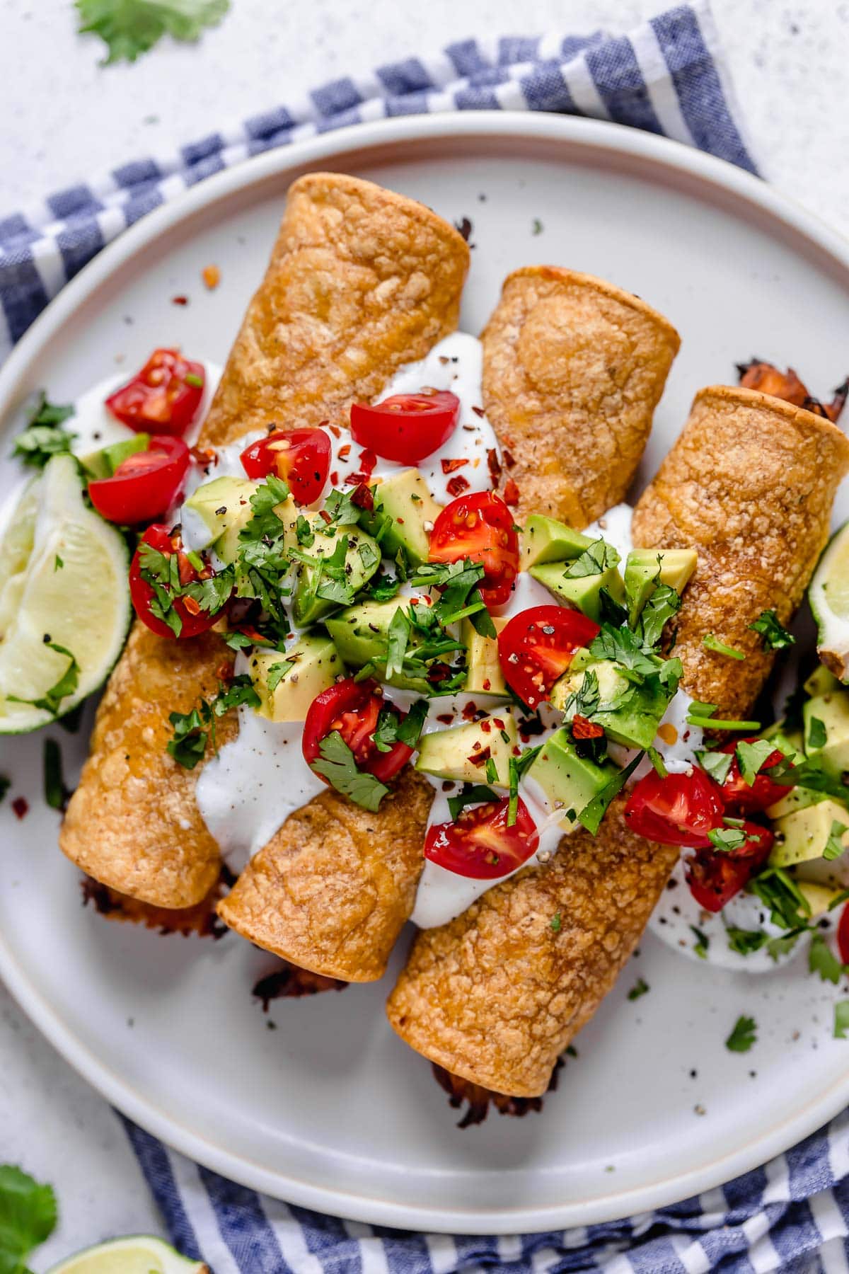 Overhead image of chicken taquitos on a white serving plate topped with sour cream, tomatoes, avocados and cilantro.