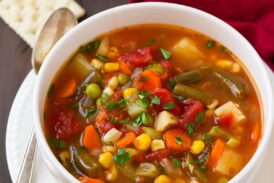 Single serving of vegetable soup in a white bowl set over a white plate. Set on a dark wooden table with a red napkin to the side.