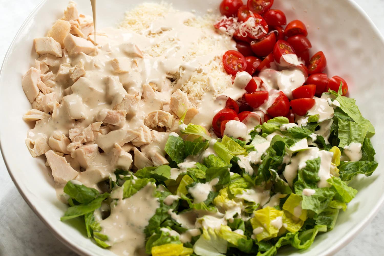 Dressing added to chicken caesar salad ingredients in mixing bowl.