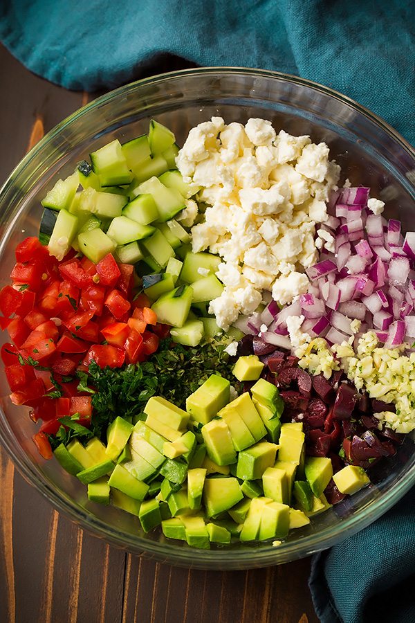 Greek salsa ingredients in a glass mixing bowl. Includes tomatoes, cucumbers, avocado, feta, red onion, garlic, olive and herbs.