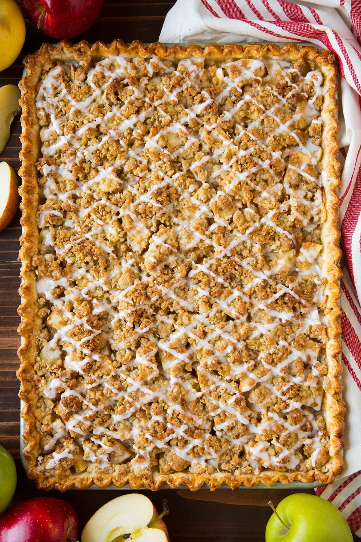 Apple slab pie with crumb topping and icing, after it has finished baking. Pie is in a rimmed sheet pan and set over a dark wooden surface.