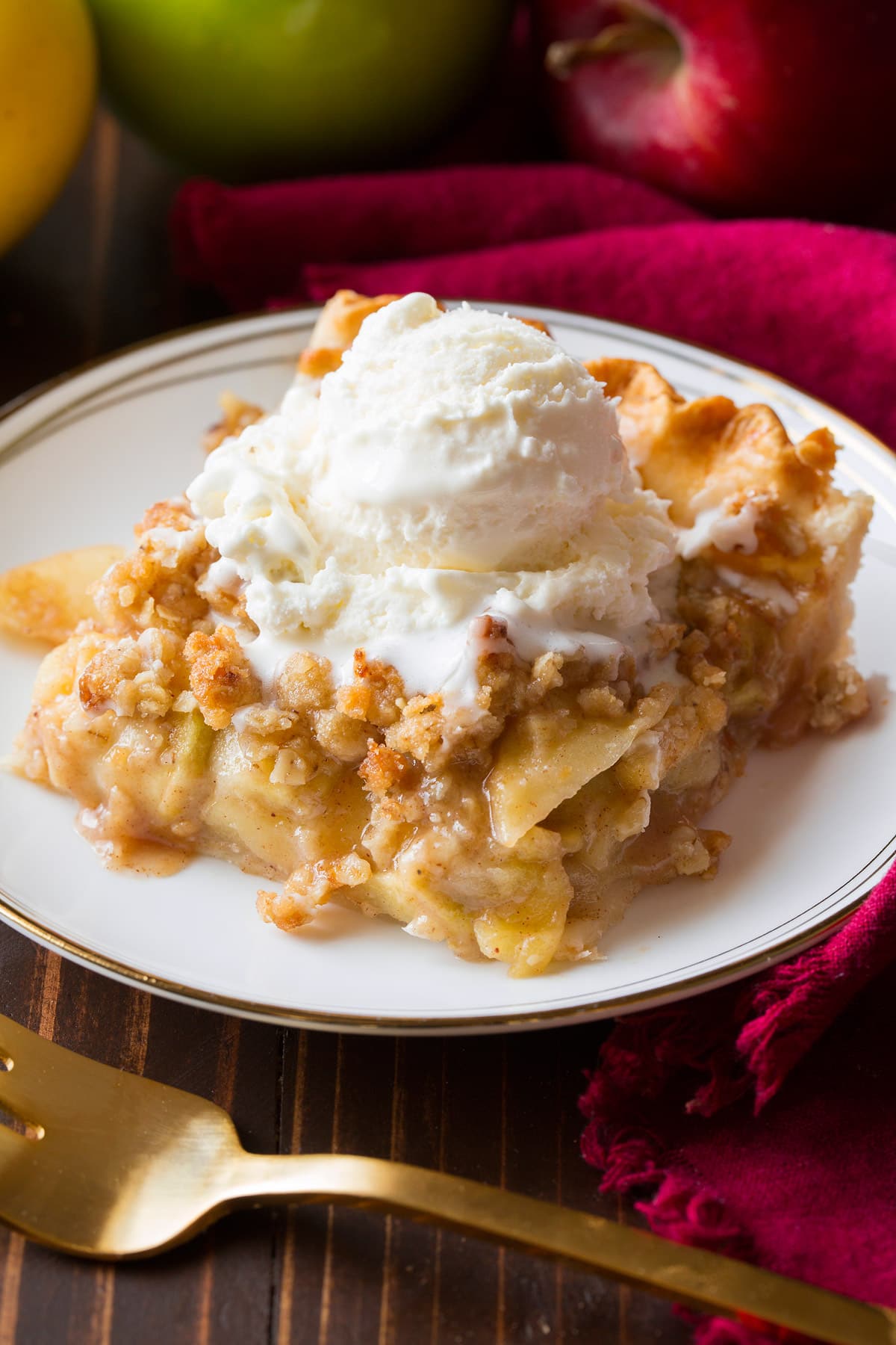 Slice of apple slab pie with crumb topping on a white dessert plate set over a brown wooden surface. Pie slice is topped with vanilla ice cream.