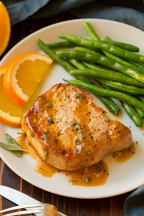 Pork chop on white plate with orange sage sauce. Served with a side of green beans and oranges slices.