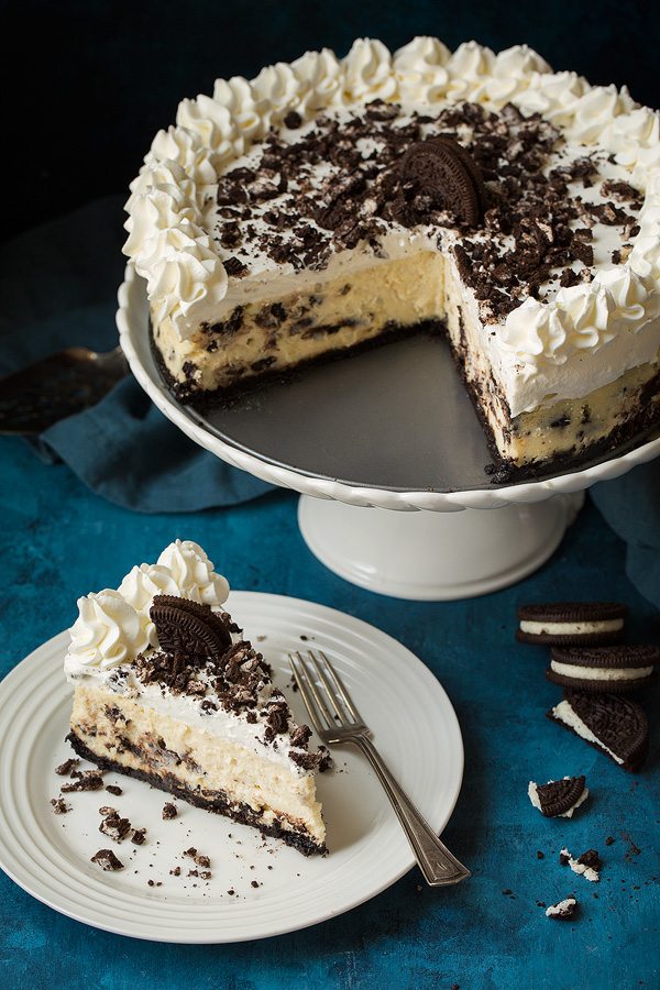 One slice of oreo cheesecake in low left corn of image. Whole cheesecake in upper right corner of image on a cake stand.