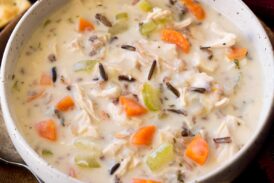 Serving of chicken and wild rice soup shown in a white bowl over a wooden plate on a black surface.