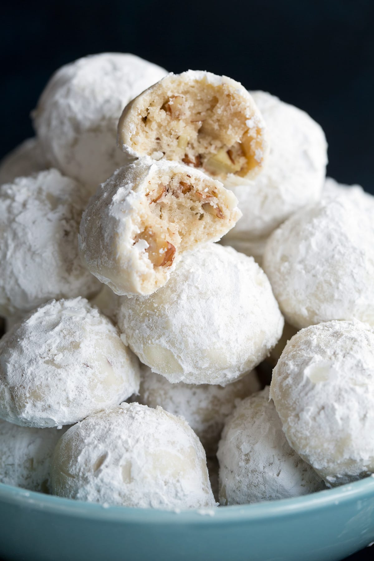 Close up image of 14 Mexican Wedding Cookies in a bowl. One broken in half to show texture.