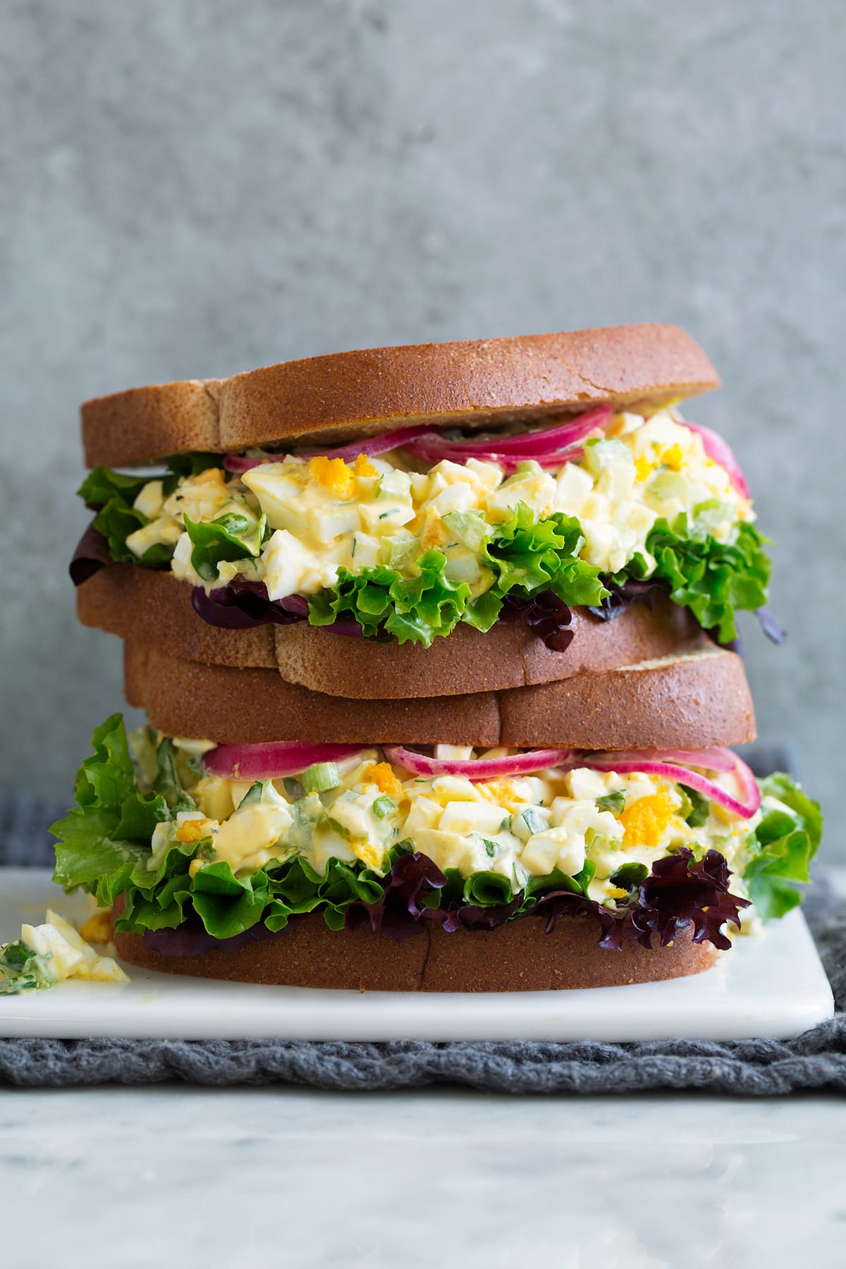 Stack of two Egg Salad Sandwiches on a white serving platter sitting on marble with a grey background.