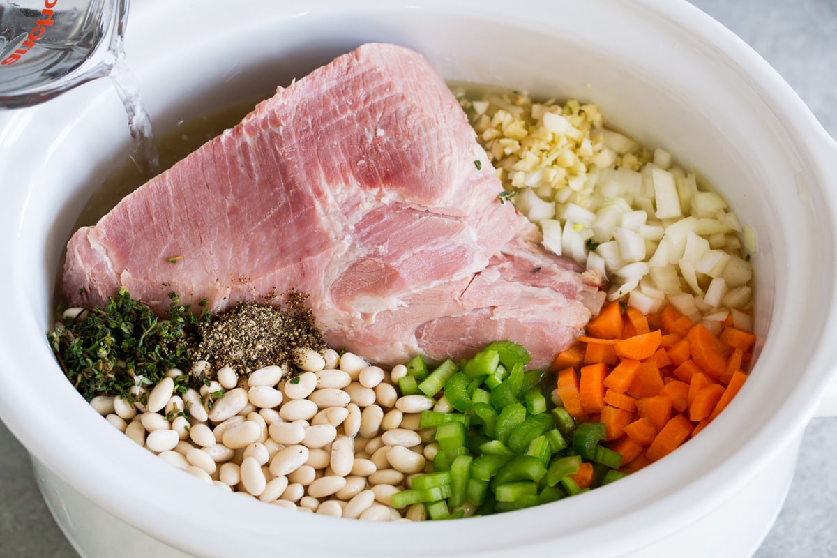 Showing how to make ham and bean soup. Adding all ingredients to a slow cooker including meaty ham bone, broth, garlic, onion, carrots, celery, great northern beans, thyme and pepper.