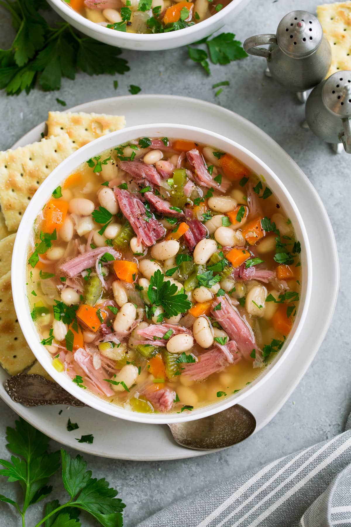 Overhead image of single serving of ham and bean soup in a white bowl set over a white plate with a side of crackers.