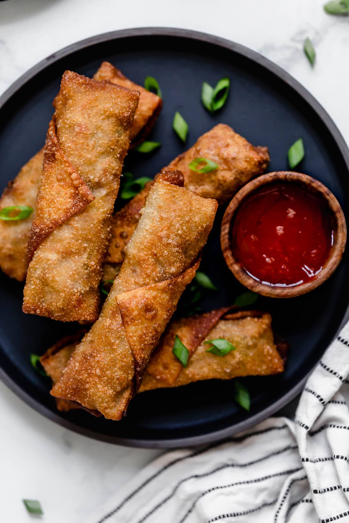 Five egg rolls on a dark plate with a side of dipping sauce.