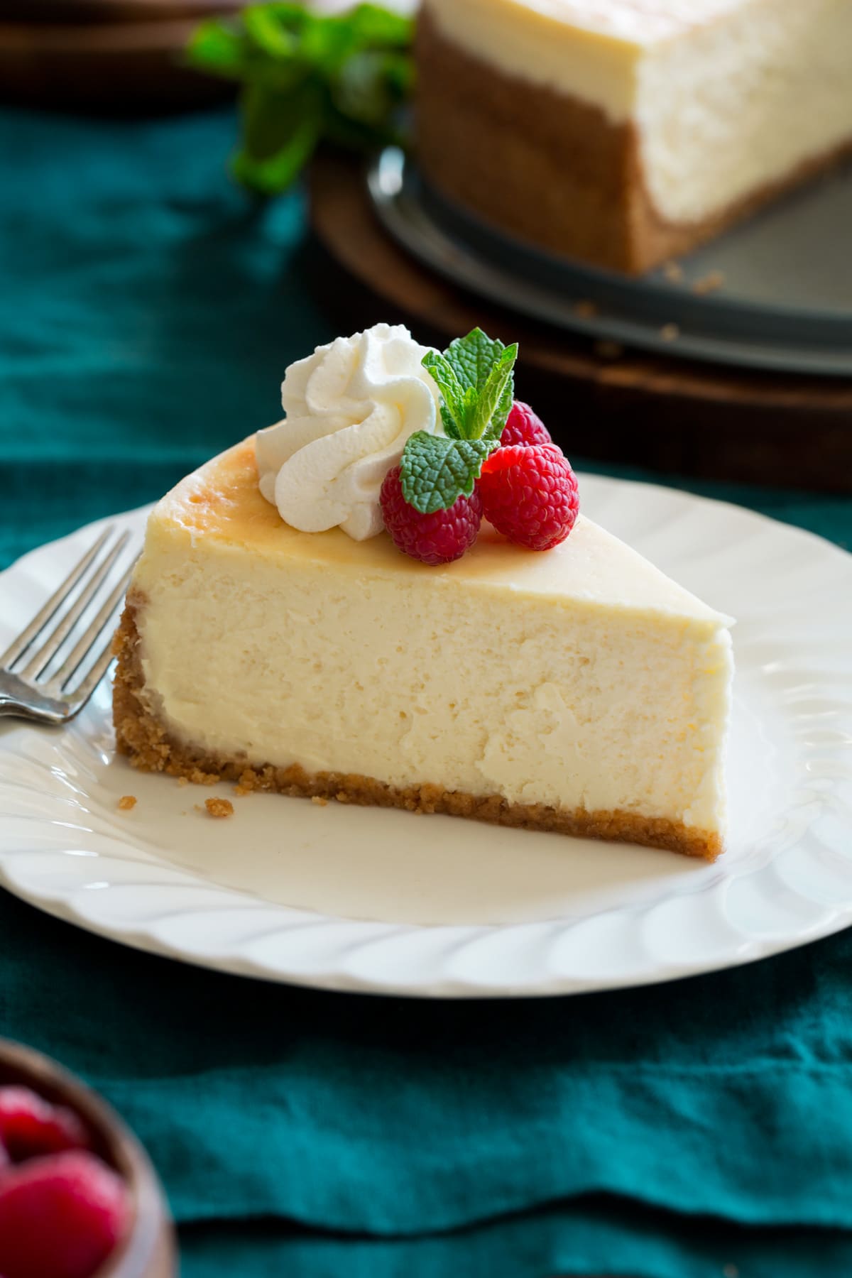 Slice of cheesecake on a white dessert plate sitting over a turquoise cloth. Cheesecake is decorated with whipped cream, raspberries and mint.