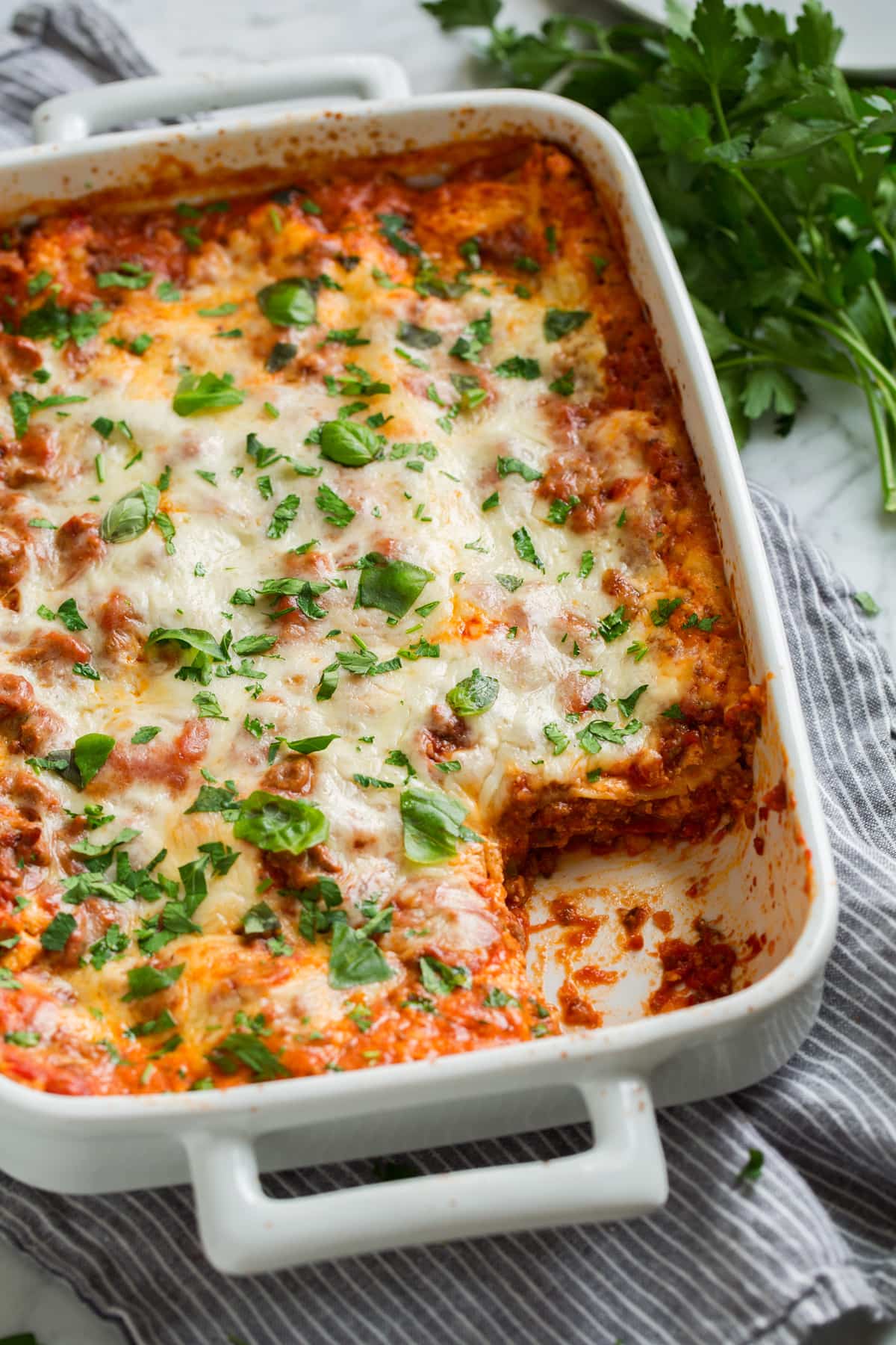 Finished lasagna in a white casserole dish with one slice scooped out. Dish shown tilted at an angle with fresh parsley in the background.