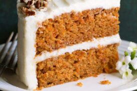 Slice of carrot cake with cream cheese frosting on a white dessert plate sitting on a green cloth napkin.