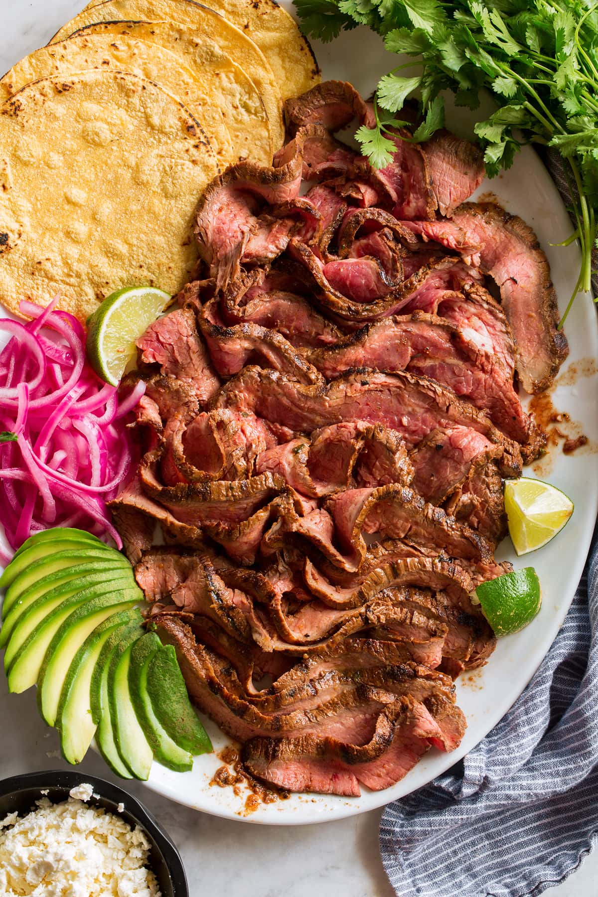 Overhead image of thin sliced carne asada on a serving platter with tortillas, onions, avocado, lime and cilantro.