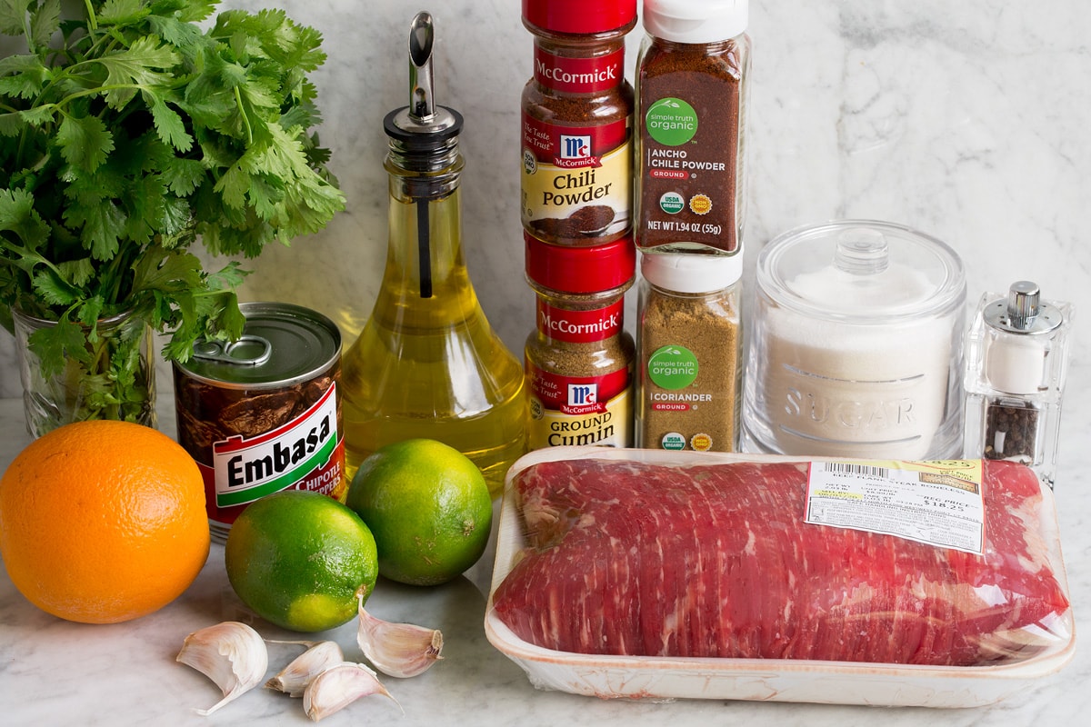 Image of ingredients used to make carne asada and marinade. Includes flank steak, cilantro, olive oil, chili powders, cumin, chipotle peppers, limes, oranges, sugar, garlic, salt and pepper.