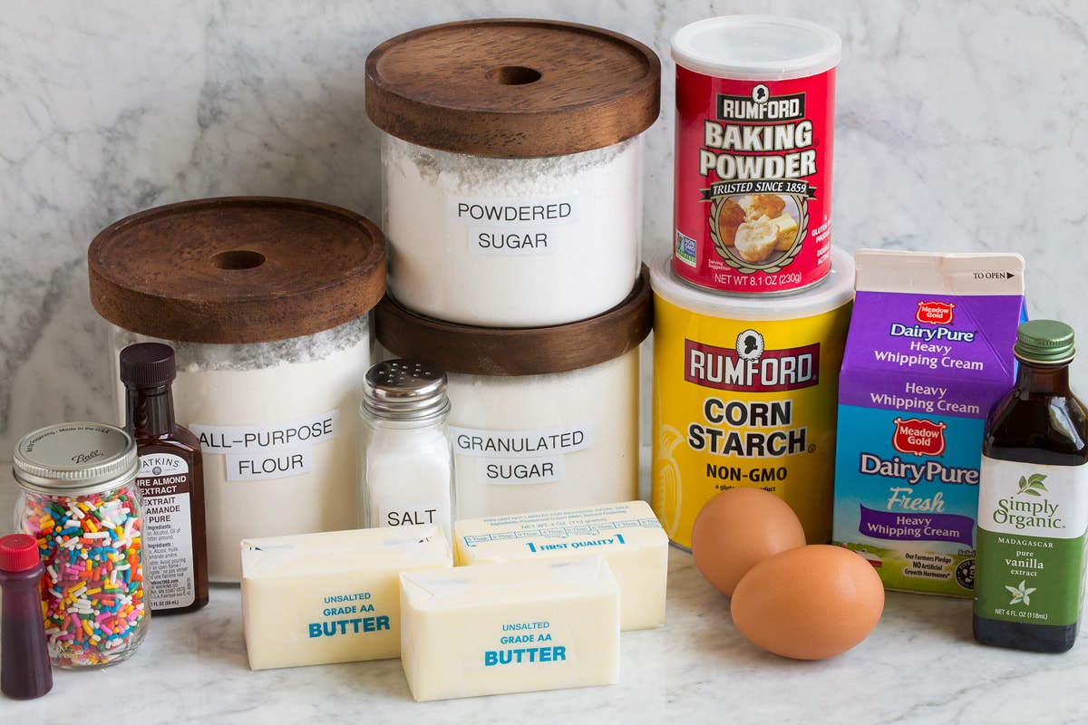 Image of ingredients that are used to make sugar cookie bars. Includes flour, sugar, powdered sugar, butter, eggs, baking powder, cornstarch, cream, vanilla, almond extract, sprinkles and food coloring.