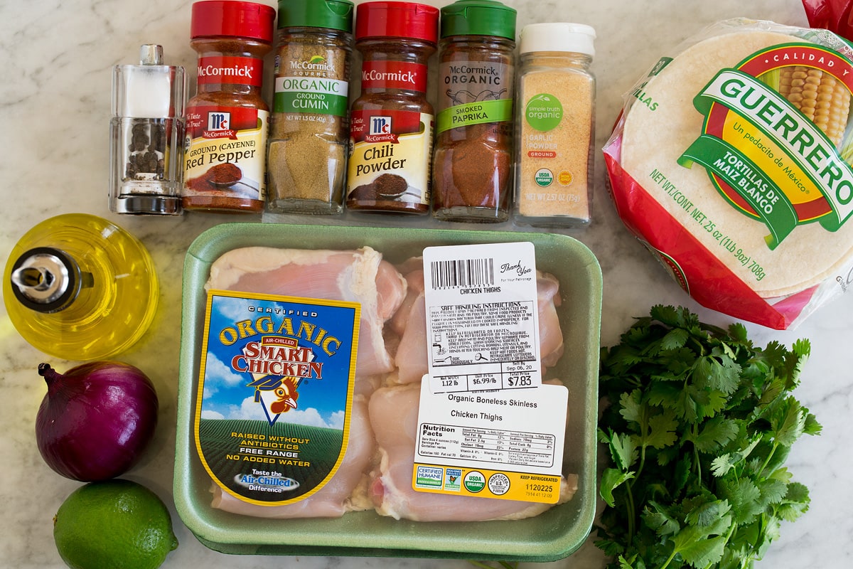 Image of ingredients used to make chicken tacos. Includes chicken thighs, chili powder, cumin, cayenne pepper, garlic powder, smokes paprika, corn tortillas, cilantro, red onion, lime and olive oil.