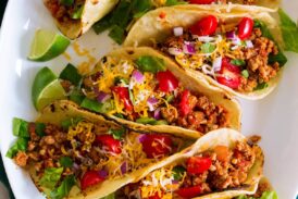 Photo: 7 ground turkey tacos in a row shown on a white oval platter from above. Tacos are filled with meat, tomatoes, lettuce, cheese and red onion.