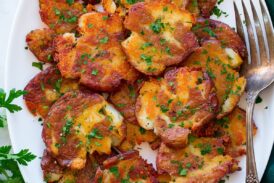 Smashed red potatoes shown piled on a white oval platter. They are shown from above on a white marble surface with a green cloth and parsley to the sides.
