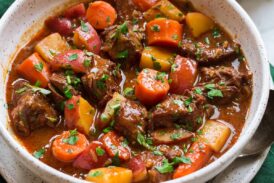 Beef Stew shown in a white serving bowl over a green cloth. Bread is shown to the side as a serving suggestion.