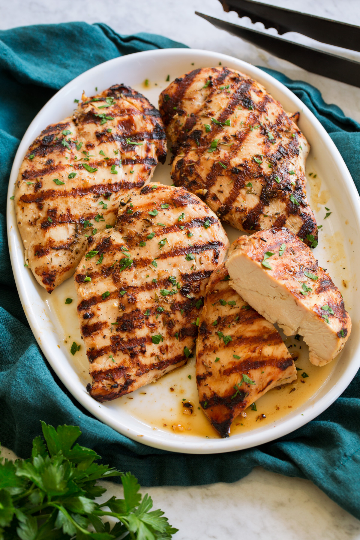 Grilled Chicken Breasts on a white platter on a teal colored cloth shown from a side angle with parsley and grilling tongs to the side.