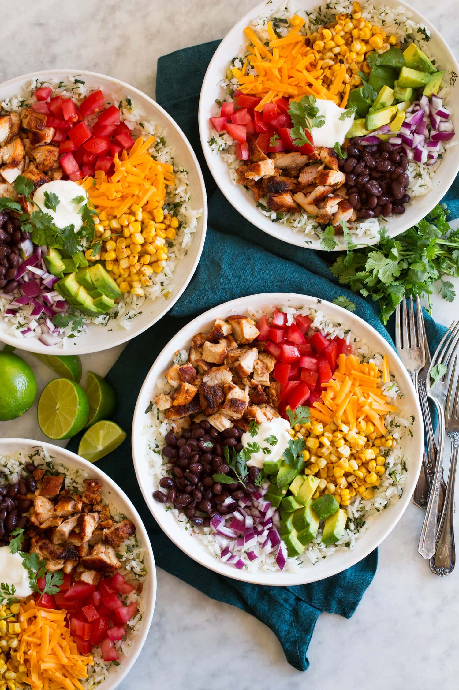 Four burrito bowl servings with corn, tomatoes, cheddar, avocado, black beans, red onion, sour cream and chopped chicken thighs.