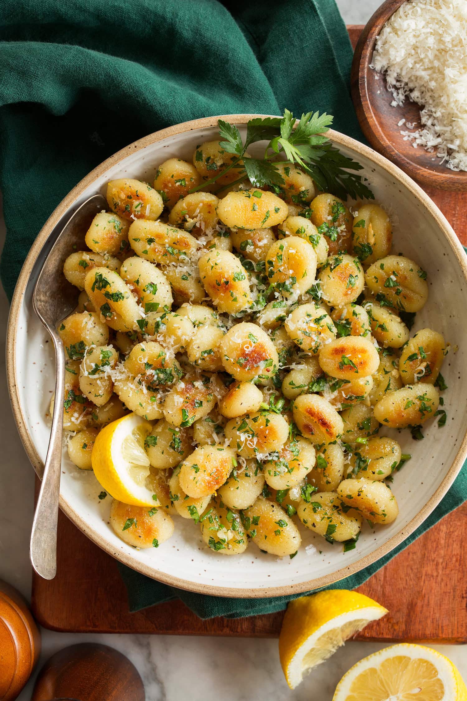 Pan fried gnocchi with lemon wedges, shown in a cream bowl over a wooden surface with a green cloth.